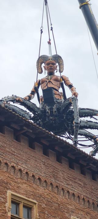 Une machine en bois et métal surmontée d'une tête de femme avec des cornes de bouc et des pattes de scorpion . Elle est sur le toit d'un bâtiment en briques et tenue par une grue