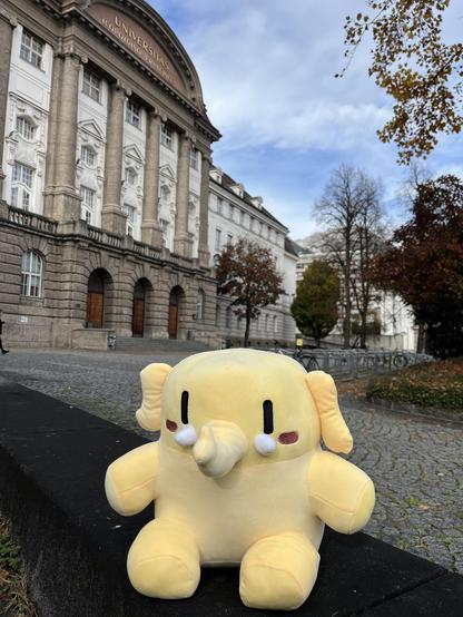 The official Mastodon stuffed toy sits in front of the main building of the University of Innsbruck, trees around, autumn scenery.