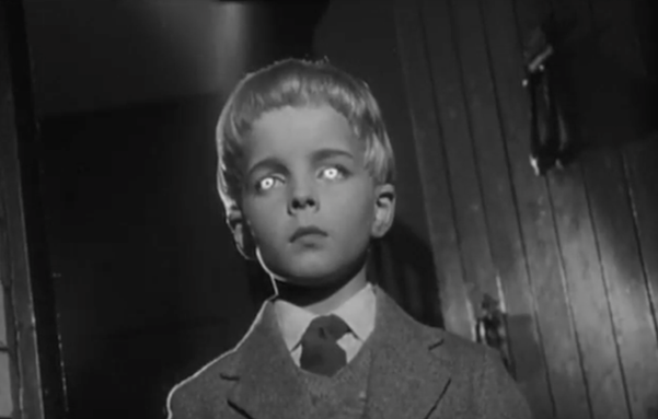 Black and white still from Village of the Damned of a young boy wearing a grey suit, white shirt, and dark tie, with short light hair almost in a bowl cut, with bright glowing eyes, in a hallway by an open wooden door with a heavy iron knocker, shot upward from below the child's eyeline at a slightly canted angle.