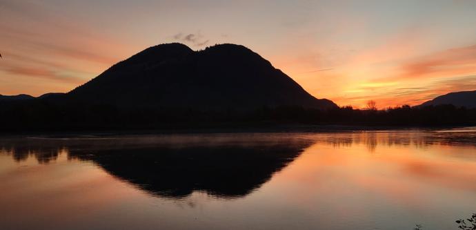 Vibrant sunrise of pink and orange sky around the silhouette of Mount Peter and Paul reflecting on the North Thompson river.