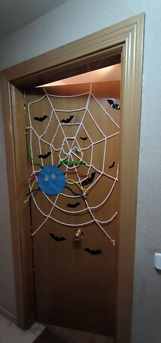 Puerta entreabierta. Una tela de araña blanca ocupa la parte media superior de la puerta. Salpicando la puerta vemos varios murciélagos de plástico.
En medio de la telaraña, un poco hacia la izquierda, una araña hecha con un círculo azul grande de fieltro, limpiapipas de colores simulando las patas y dos ojos blancos con muchas pupilas negras.