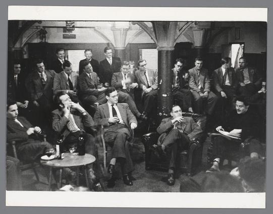 The image is a black and white photograph depicting a gathering of people, likely in the mid-20th century judging by their attire. It appears to be taken indoors at an event or meeting held on Leidseplein, Amsterdam, as indicated by Lucebert's work displayed above them. The individuals are engaged in various activities such as conversing, reading, and observing something out of frame.

The men are mostly dressed in suits with ties, while the women wear dresses and coats typical of that era. Some people sit on chairs or benches arranged around small round tables, which have items like glasses and papers placed upon them. The setting suggests a social club or student association gathering where intellectuals might be discussing topics related to their interests.

The atmosphere is somewhat casual yet attentive; some are deep in thought while others converse with one another, indicating an informal but engaged environment.