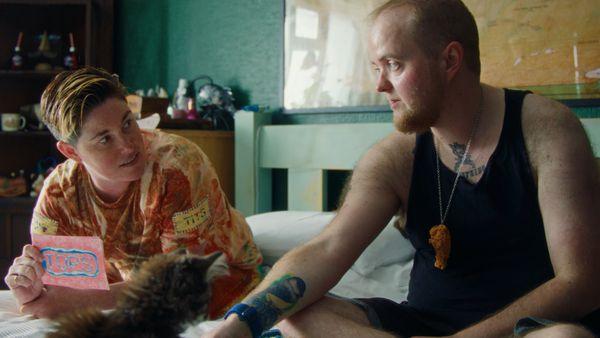A still from the film Tops. Two white trans men sit on a bed along with a scruffy-looking tawny kitten. One of them is wearing a dark vest top and shorts and looking relaxed but interested. The other is wearing a yellow and orange t-shirt and looking anxious.