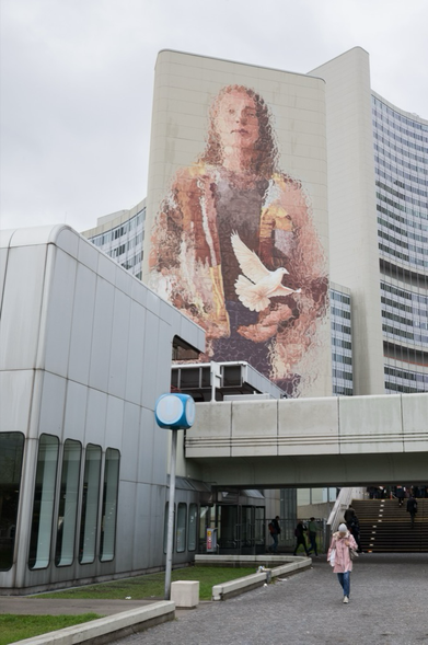 A tall building features a mural of a person holding a dove, with warm pink and brown tones. Below, a woman walks under an overpass wearing a pink coat, matching the colors in the mural. The scene captures a striking harmony between the mural and the woman's outfit against the backdrop of the modern, curved architecture and overcast sky.