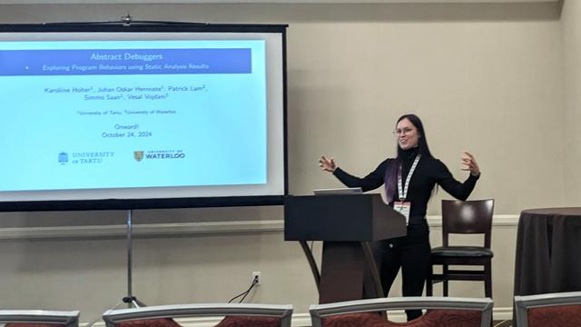 A person at a podium making an explanatory gesture, with the title slide ("Abstract Debuggers: Exploring Program Behaviors using Static Analysis Results") being projected behind.