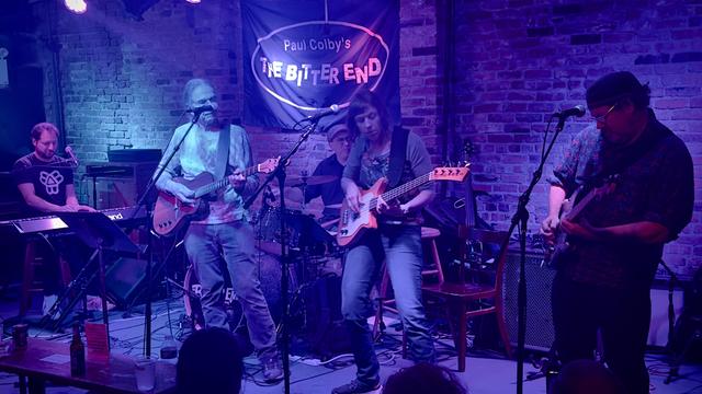 David Gans and the Broken Angels playing The Bitter End on Bleeker Street in Greenwich Village NYC on Thursday October 24, 2024. The band is playing on stage, which is in front of a brick wall and lit with purple, red & blue lights.