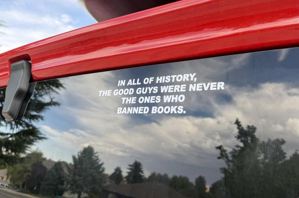 A white-lettered sticker on tinted rear window of a red vehicle reads: "IN ALL OF HISTORY, THE GOOD GUYS WERE NEVER THE ONES WHO BANNED BOOKS."  Reflected sky is a bonus.