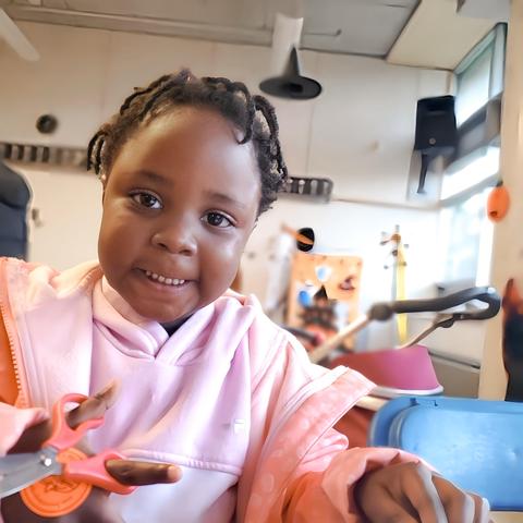A young black girl with little braids, holding scissors in her right hand, wearing a pink hoodie & peach jacket, smiling at the camera.