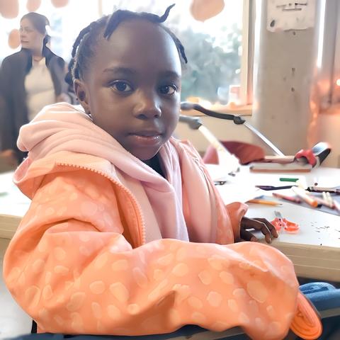A young black girl, with some small dreadlocks, wearing a pink hoodie & peach jacket, sitting sideways beside arts/crafts table & looking at camera with small shy smile.