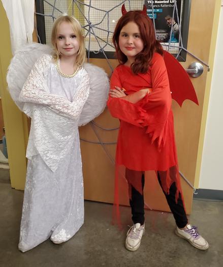 A blonde girl in white good angel costume & a redhead girl in red & black bad angel costume. Standing in front if a door covered in corded spiderweb.