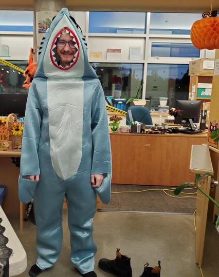 A bearded man with glasses in a full great white shark costume. Standing in front of office reception desks.