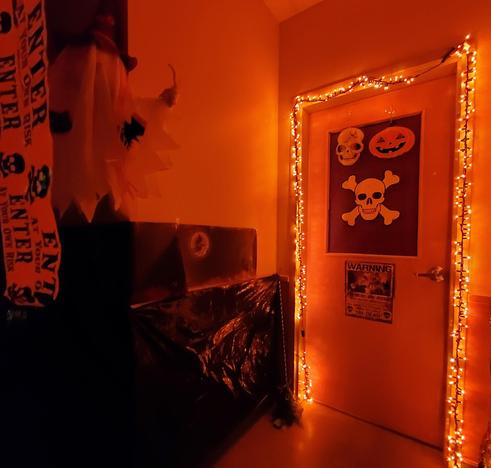 Part of the haunted house area with orange lighting, skulls & jack o lantern on a door with zombie warning sign, a ghost hanging from the top upper left corner of ceiling with enter at your own risk tape streaming from ceiling.