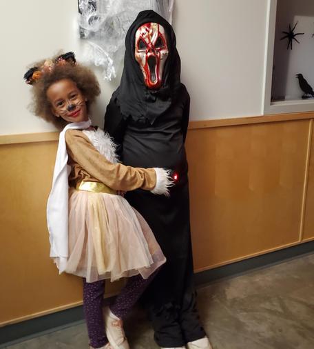 A black girl in a Halloween cat costume with painted whiskers & nose. Hugging her friend, dressed as Death. They're standing against a wall in hallway.