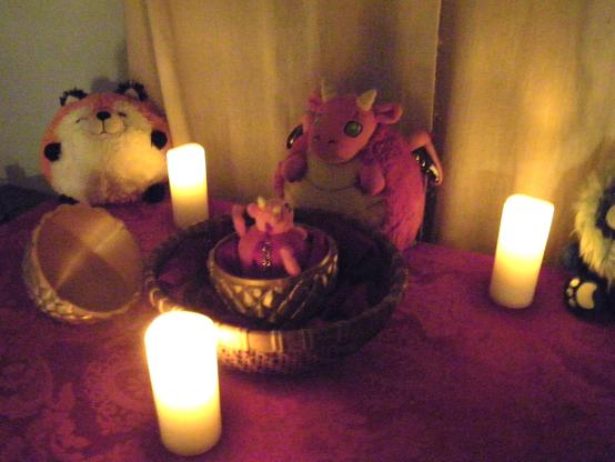 Three plush toys, surrounding a wicker basket containing a golden, dragon-scaled egg that has opened. Inside the egg is a baby red and yellow dragon. There are three candles surrounding the basket, and a red altar cloth. The plush toys are an orange and white fox with black feet, a red and yellow dragon with green eyes, and a blue lion with a yellow moon symbol and black wings. The red dragon is very excited to see the baby dragon.