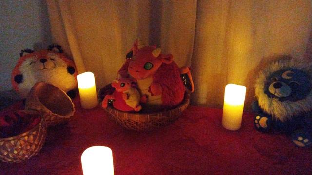 Two plush toys, surrounding a wicker basket containing a mama and baby red and yellow dragon plush toys. There are three candles surrounding the basket, and a red altar cloth. The plush toys are an orange and white fox with black feet, two red and yellow dragons with green eyes and gold wings, and a blue lion with a yellow moon symbol and black wings. The red dragon is holding the baby dragon.