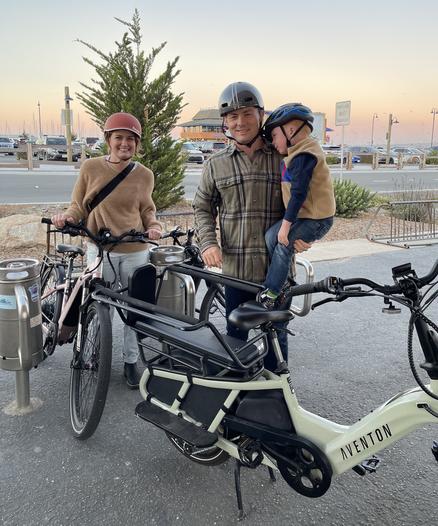 A woman and man—and in his arms, their young child—stand alongside the two bicycles they are about to ride. Both are Aventon ebikes, and the one the father rides has a child carrier on the back.