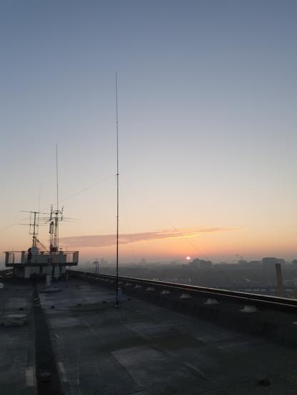 Auf einem schmalen Dach steht eine vertikale Antenne. Im Hintergrund geht im Nebel die Sonne auf.
Am hinteren Ende des Dachs ist eine Aussichtsplattform und weitere Antennen zu sehen.