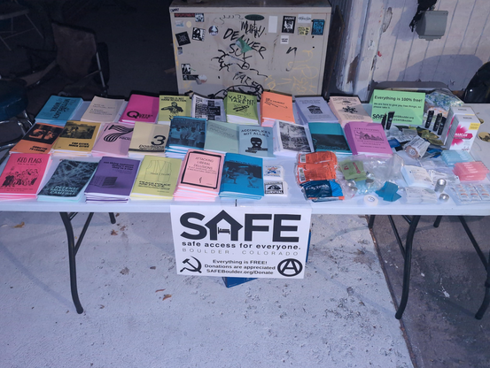 a folding table covered in stacks of zines in different colors, with harm reduction supplies on one corner, including boxes of Narcan.

A sign with the SAFE logo is taped to the front of the table.