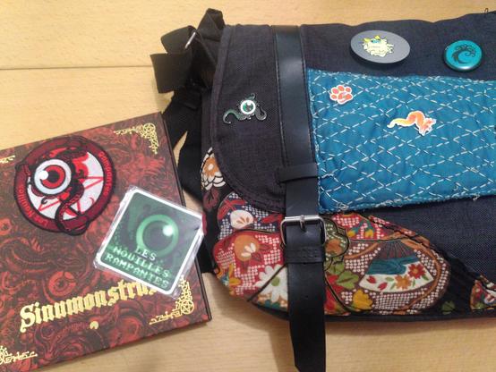 Sur une table, un livre rouge et doré, avec un écusson de tissu et un magnet, représentant tous deux un œil avec des tentacules. À côté, un sac bandoulière avec le rabat couvert de pin's : le même œil, une trace de patte de chien, un renard de feu, un chat touffu, un arbre bleu-vert.