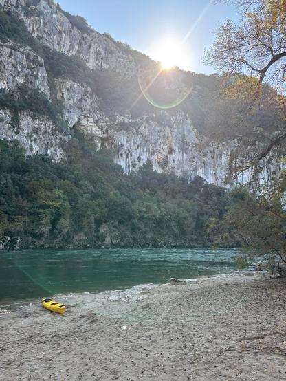 A landscape with a turquoise-coloured river winding through a valley. The sun is shining over a steep cliff which is covered in plants. The other side is lined with trees in autumny colours. A yellow kayak sits on the sandy shore.