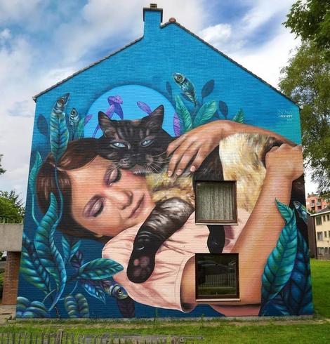 Colorful mural titled 'Jade and Moggy' by artist Nina Valkhoff in Gent, Belgium, showing a serene girl embracing a black cat. The scene is surrounded by vibrant blue and green leaves with small, whimsical fish appearing around them. The mural captures a tender moment, symbolizing love and comfort, based on a photograph by Karin Post of her daughter Jade and their cat Moggy. This artwork, created for the Goldmine festival organized by Wallin’, aims to convey a message of hope and the universal need for a comforting hug.
