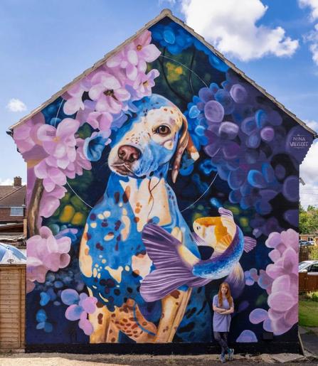 Large mural by artist Nina Valkhoff in Cheltenham, Gloucestershire, created for the Cheltenham Paint Festival. The artwork depicts a Dalmatian dog surrounded by vibrant purple and pink flowers with a large, colorful fish swimming nearby. The intricate details and vibrant colors make the scene dreamlike, capturing a surreal interaction between the dog and fish. The artist, standing beside the mural, adds a sense of scale to this whimsical, nature-inspired piece.