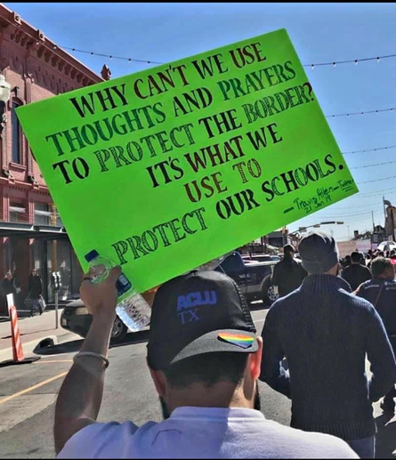 a man walking in the street, participating in a march is holding a sign that reads:
"Why can't we use thoughts and prayers to protect the border? it's what we use to protect our schools."
