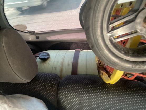 Das Bild zeigt den Innenraum eines Fahrzeugs, mit einer teilweise sichtbaren Gasflasche, die mit einem Gurt befestigt ist, zusammen mit einem Rad und einem Teil eines Sitzes. Der Hintergrund zeigt unscharfen Straßenverkehr an.