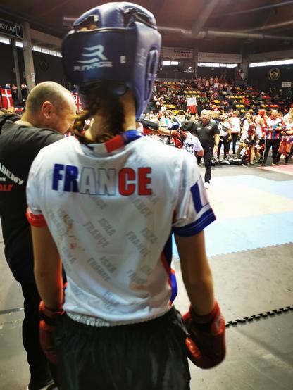 Silhouette de boxeuse vue de dos, s'apprêtant à monter sur le tatamis. Sur son t-shirt apparaît le mot France. Elle porte un casque et des gants.