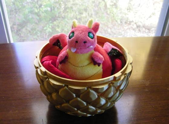 A baby dragon plush sitting in a golden dragon egg. The dragon plush is mostly red, with a yellow belly and horns, green eyes, and wings with gold highlights. The dragon is on a wooden table, and in the background is a window.