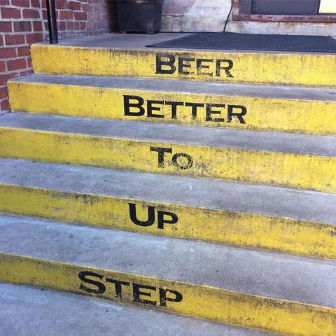 Five concrete steps lead up to a concrete platform with a black mat on it.  The front of each step has a word written in black, highlighted in yellow.  The five words are Step Up To Better Beer.  The bottom of a red brick wall can also be seen to the left of the steps.