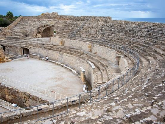 Blick auf das römische Amphitheater in Tarragona. Man sieht die Sitzränge aus Stein, dahinter ist der blaue Himmel zu sehen.