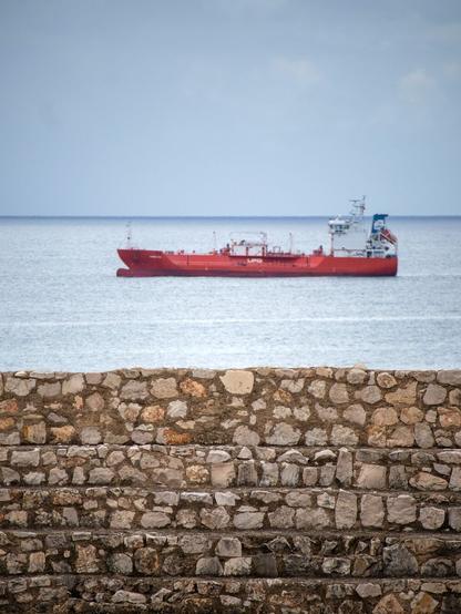 Ein Blick über die Gemäuer des Amphitheaters. Über dem steinernen Bauwerk ist das Meer zu sehen und parallel dazu ein rotes Frachtschiff.