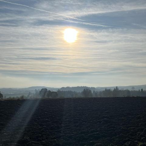 Square photo of an autumn afternoon. There is a brown plowed field in the foreground, trees and gentle misty hills in the background. The golden sun is shining in a blue sky with cirrostrati.