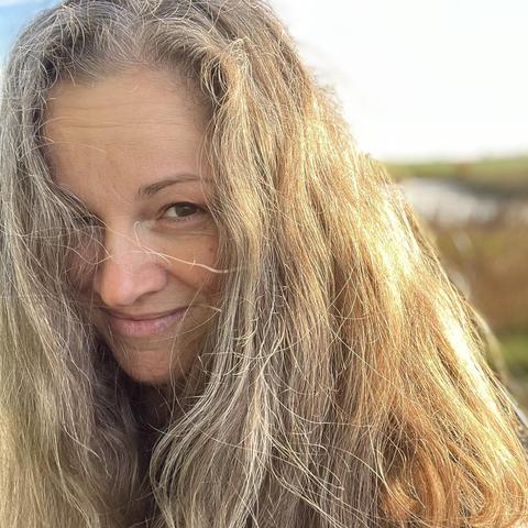 Square photo of Abyss Cannonball’s head with the golden October sun shining upon her from the right. She has long brown and gray hair, brown eyes, and she is smiling at you. She looks happy about the lovely autumn weather.
