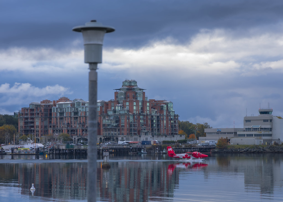 A red and white seaplane (Canadian flag colours and maple leaf graphics, kind of) slowly cuts through calm water. It is reflected in the water. Behind it, multistory buildings, perhaps an apartment building, look on & also reflect on the water. In the foreground, an out of focus streetlamp occupies the left side of the image.