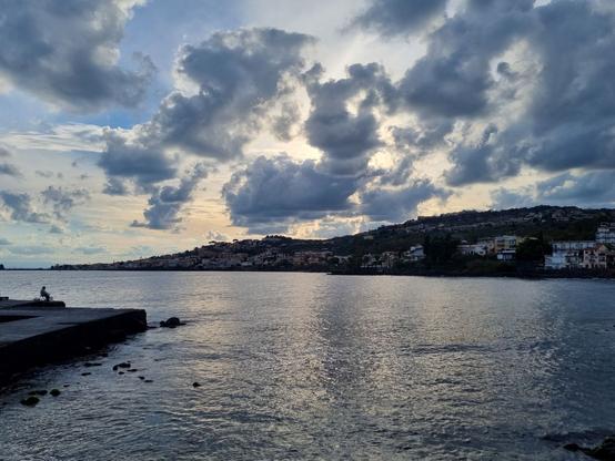Sole al tramonto, nascosto dalle nuvole, riflessi di luce sul mare calmo e anziano pescatore seduto con una canna da pesca in mano