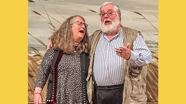 Photograph of Anni Fentiman on the left and Dave Webber on the right, performing at the Everyman Folk Club on Friday 25 October. Anni is wearing a black and white dress with a black scarf. Dave is wearing a striped shirt and brown jacket. Dave's arm is around Anni's shoulders, and they are both singing.