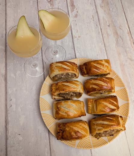 A photo taken from above of two cocktails and a plate with eight sausage rolls on it, sitting on a white wooden background. The sausage rolls are golden brown, with three small scores in top of each, and a brown sausage filling. The cocktails are a cloudy apple-juice coloured drink in Nick and Nora glasses with a thin pear slice garnish each.