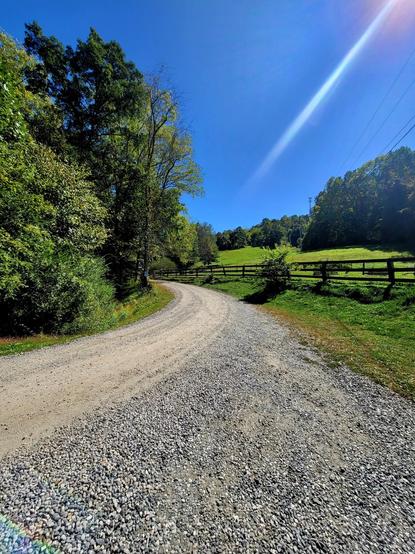 Color Photo
Very blue sky with a sun ray. 
Green field with a dark wood fence. 
Gravel drive cuts through the middle of the photo. 
Full leave trees in different shades of green on the left side.