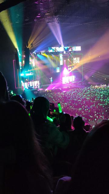 Photo from Miku Expo 2024 in London. There are a lot of people in an auditorium waving colourful pen lights. Most of the pen lights are set to green, making it seem like they are waving leeks around. Everyone is looking at a diminutive figure on a stage, surrounded by fancy lighting effects and (if you look really closely) a live band. The figure has long teal hair, oo-ee-oo...