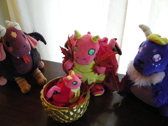 Four plush toys on a wooden table. Three of them are surrounding a half of a golden, scaled egg which contains a baby red and yellow dragon with green eyes. There are red and yellow leaves in the egg/nest. To the left of the egg is a reddish brown creature with white horns and chicken feet. In the center is a red and yellow dragon with green eyes, holding two leaves. To the right is a purple sheep/goat-like creature with a light pink beard, and holding a leaf.