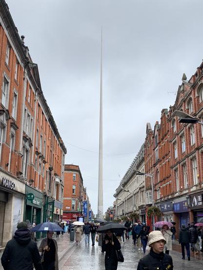 Street view of Dublin’s O’Connell Street on a rainy day, featuring the Spire of Dublin in the center. The Spire, a tall, slender stainless steel monument, stretches up into a cloudy gray sky. People with umbrellas walk along the wet, reflective pavement lined with historic red-brick and stone buildings. Shopfronts and signs add color to the scene, contrasting with the subdued, overcast atmosphere.