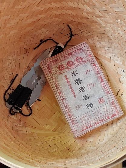 The discovery: From a point of view of staring down a bamboo basket constructed to store tea cakes can be seen three cheap tea knives and, resting atop them, a 250g brick of tea wrapped in crepe paper.  The paper has writing in Chinese both in black and in red.  (A later photograph in the sequence will hold the translation.)

This was the surprise that greeted me when I took down the basket to dust it off.