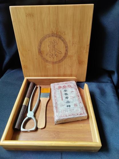 The tray is removed from the box and placed in front (horizontally, as it would usually be stored) with the box still standing vertically in the background for display purposes.  The box contains from left to right a wooden dowel with a dual brass ring in the middle that looks like it should be separable there, a strange-looking pair of pliers with two sharp "teeth" on each side, and ordinary looking paintbrush like the kind you can find for cheap in any hardware store, and the tea brick, still wrapped, from the basket photograph.