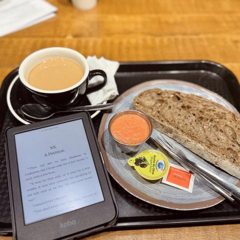 Bandeja con todo lo necesario para desayunar: café rico con leche de avena, tostada de tomate y aceite, y un libro (electrónico).