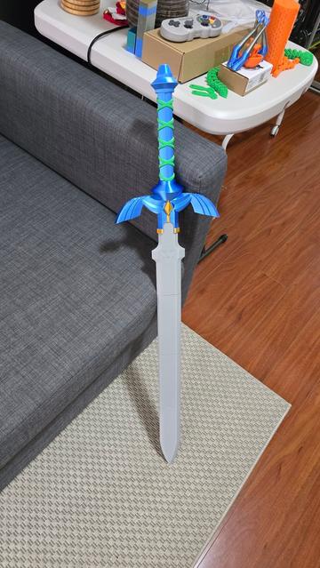 A 3D printed Master Sword in full colour with a shiny blue handle, a gold piece in the middle of the handle and green wrap around the top. It's sitting against a couch.