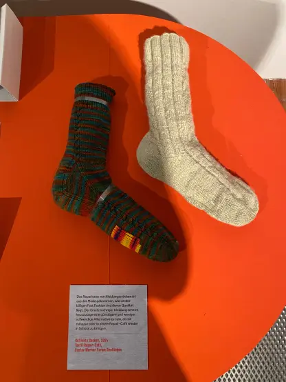 An orange table top form above.
There are two socks mounted on it with clear plastic strips.
On the left a sock knitted out of dark teal and dark orange striping yarn. A small cable runs on the side of the sock all the way from the toe to the cuff. There's a patch of brightly coloured (yellow, red, purple) yarn on the sole.
On the right a sock out of fuzzy pale green yarn. Cables and run all around the legt and on top of the foot. There are patches on the sole, but this itme in the same yarn. That's why they are almost invisible.
A plaque next to the socks reads (translated from German):
"Repairing garments has gone out of fashion, which is due to the cheap fast fashion and its quality. The replacement of clothes with holes seems to be a cheaper and less labour intensive alternative nowadays, than to repair them at home or in a repair café."
These socks were knitted many years ago and repaired this year in a repair café by me.