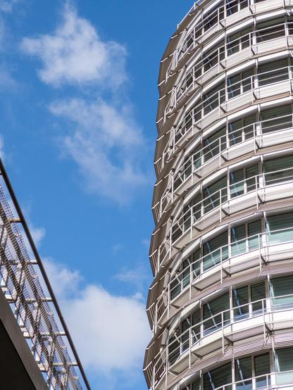 The image captures a modern, curved building with an intricate metal and glass facade under a bright blue sky. Adjacent to the structure, a section of metal railing extends into the frame, creating a dynamic architectural contrast. The mood is vibrant and contemporary.