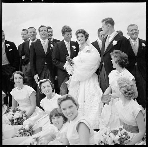 A black and white photograph capturing a joyous moment during what appears to be an outdoor wedding ceremony. The image displays several individuals dressed in formal attire, including suits for the men and dresses or gowns with flower accessories for women. At the center stands a bride wearing a bridal gown adorned with floral patterns, holding a bouquet of flowers, while being surrounded by members of her wedding party who are all smiling and looking off to the side. The group is assembled on what seems like an open terrace or balcony under overcast skies.